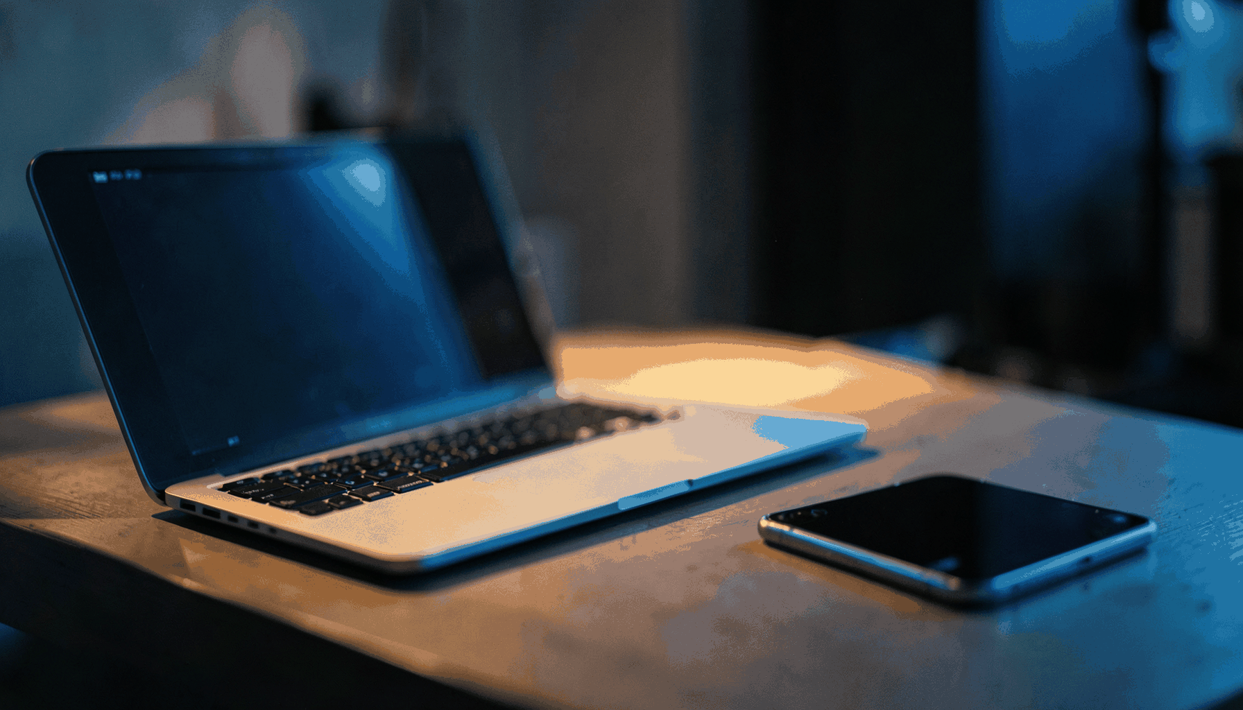 A softly lit desk scene with a laptop, notebook, and smartphone, warm amber and deep indigo tones, shallow depth of field, cinematic wide frame